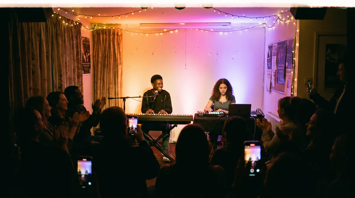 A teenager plays keyboard and another runs a beat from a laptop on stage at a student showcase while families film on their phones